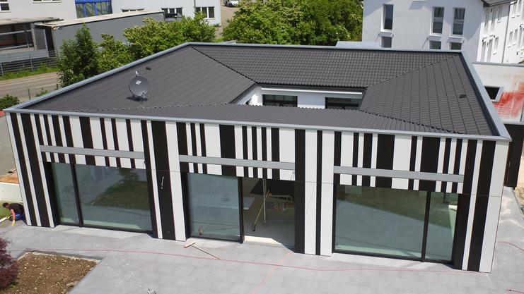 Moderne, rechteckige Gebäudeansicht mit schwarz-weißer Streifenfassade und großen Fenstern, umgeben von Rasen und Gehweg.