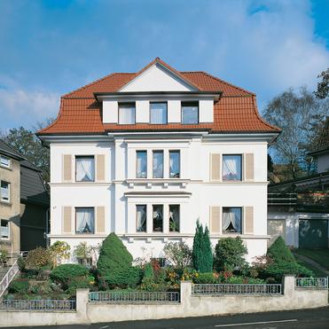 Zweifamilienhaus mit rotem Satteldach, weißen Wänden und Garten, umgeben von weiteren Gebäuden unter blauem Himmel.