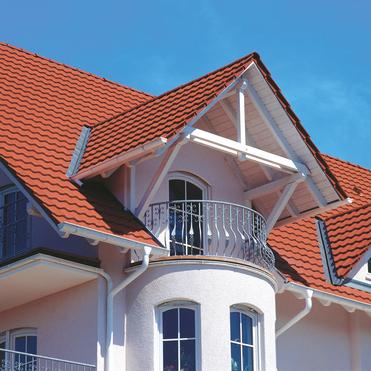 Detailansicht eines Hauses mit roter Dachdeckung und Balkon, unter blauem Himmel.