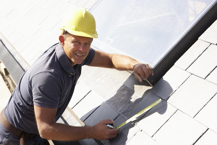 Handwerker mit gelbem Helm misst die Länge von Dachziegeln auf einem geneigten Dach. Sonnenlicht reflektiert auf Solarpanel.