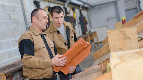 Zwei Männer in Arbeitskleidung betrachten Ziegelsteine an einem Tisch in einer Werkstatt.